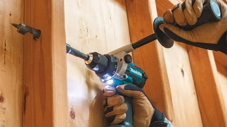 A person in protective gloves is operating a Makita cordless hammer drill with an illuminated LED work light, drilling into a wooden surface, with a focus on precision and safety in a woodworking environment.