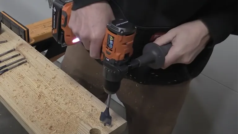 A close-up of a person using a RIDGID hammer drill with an illuminated LED light, drilling into a plank of wood with drill bits visible on the side.