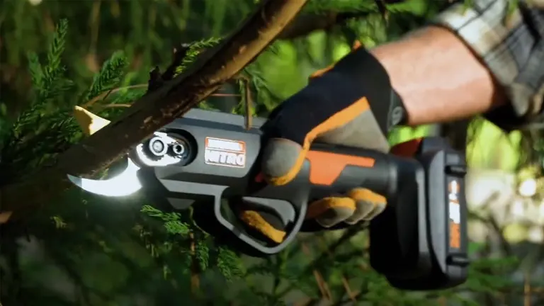 A close-up of a gloved hand using a Worx NITRO electric pruning shear to cut a branch among green foliage.