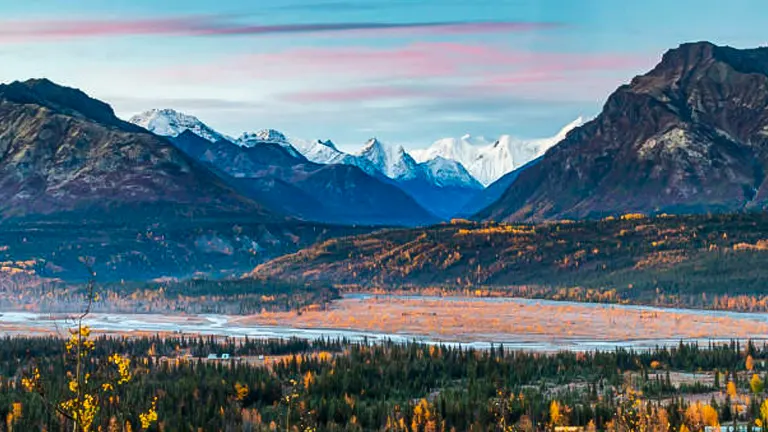 Autumn colors blanket the landscape of Chugach National Forest, with snow-capped mountains in the distance under a dawn sky with pink-hued clouds.