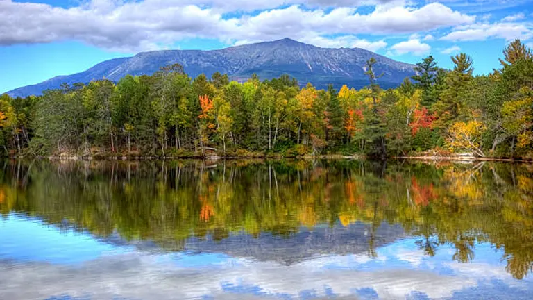 Reflective lake with autumnal foliage in the foreground and a large mountain under a cloudy sky in the background.