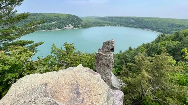 Overlook of a serene turquoise lake from a rocky cliff with lush green trees under a clear sky with wispy clouds.