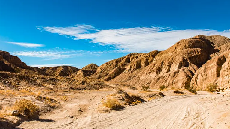 A dusty trail leads through a dry, rugged desert landscape with undulating hills under a clear blue sky.