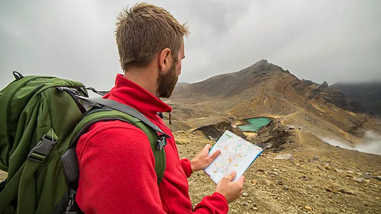 A bearded hiker in a red jacket with a green backpack consulting a map in a mountainous landscape with misty skies and a glimpse of emerald lakes in the distance.