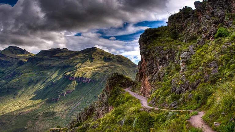 A winding trail along a mountainous landscape with lush greenery, under a dramatic sky with billowing clouds.