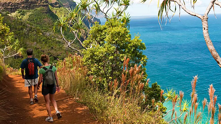 A couple hiking on a coastal trail with lush greenery, overlooking the deep blue sea, with a mountainous backdrop and clear skies.