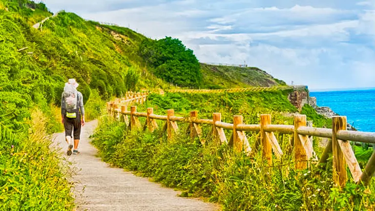 A lone hiker walking on a seaside path with wooden railings, green hillside to one side and the ocean to the other, under a partly cloudy sky.
