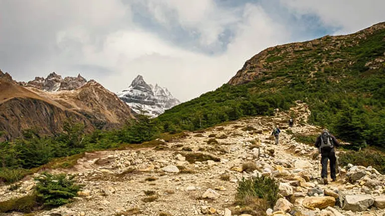 A lone hiker trekking a rocky path in a mountainous terrain, with sharp peaks in the background partially shrouded by clouds.