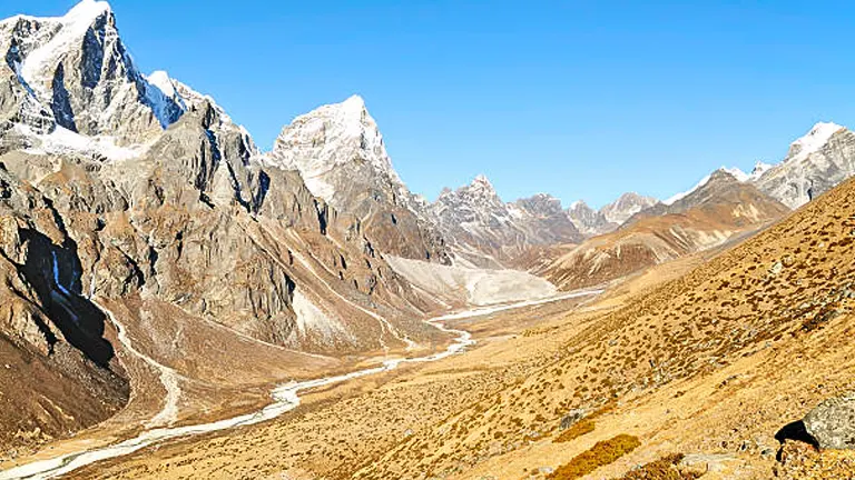 A picturesque view of a winding river valley flanked by rugged, snow-capped mountains under a clear blue sky with a hiker visible in the foreground.