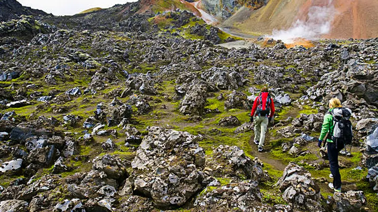 Two hikers navigating a rugged lava field with green moss, with steam rising from geothermal vents near colorful rhyolite mountains in the background.