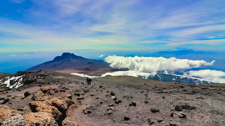 A hiker on a volcanic terrain with sparse snow patches, facing a distant mountain peak above the clouds, under a gradient blue sky.