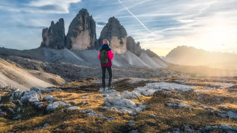 A solitary hiker in a red jacket standing in a rocky alpine landscape, gazing at the towering jagged peaks of the Tre Cime di Lavaredo at sunset.