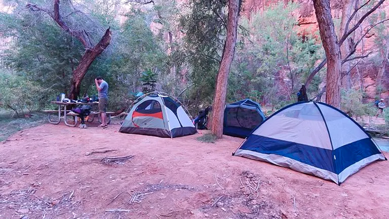 A camping setup with two tents and a camper attending to gear on a picnic table, surrounded by trees with towering red cliffs in the background, creating a serene outdoor living space.
