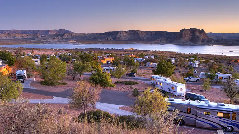 An RV park at dusk with numerous campers and RVs set against a backdrop of Lake Powell and its surrounding cliffs, under a twilight sky.

