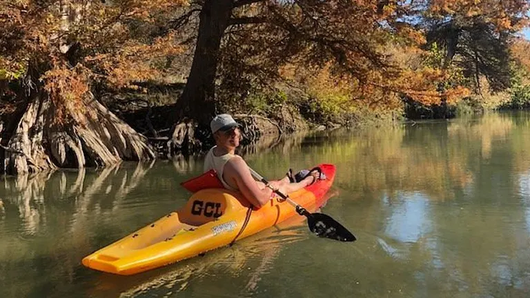 A person in a yellow kayak enjoying a leisurely paddle on a calm river, flanked by trees with autumn foliage.