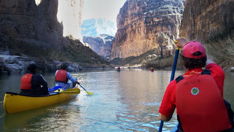 Kayakers in a calm river canyon with steep, rugged cliffs rising on either side, under a clear sky. The distant walls are touched by sunlight, highlighting their textures and the expansive scale of the canyon.