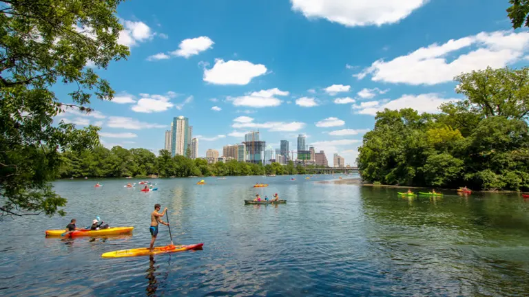 People engaged in various water activities including kayaking and stand-up paddleboarding on a tranquil river with a backdrop of a bustling city skyline and lush greenery under a blue sky with fluffy clouds.