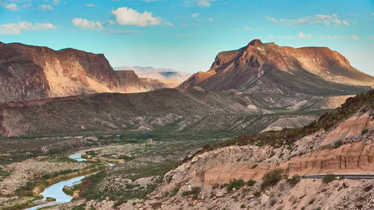A panoramic view of a rugged desert canyon with a river snaking through it, a road hugging its side, and mountains rising in the background under a dusky sky.

