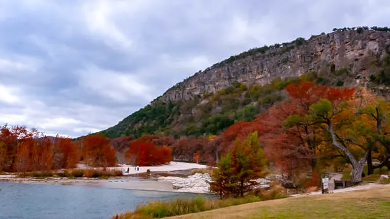 An overcast sky looms over a tranquil river flanked by trees with autumn foliage and a towering limestone cliff in the background.
