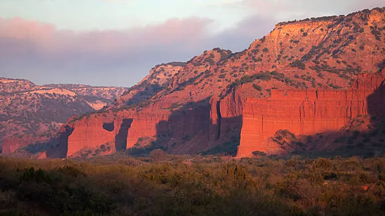Reddish cliffs basked in the warm glow of sunrise or sunset, with the foreground of lush greenery under a soft, dusky sky.

