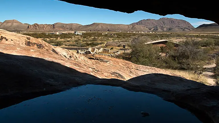 A view from a cave opening overlooking a desert landscape with rocky ground, sparse vegetation, and mountainous backdrop under a clear blue sky.
