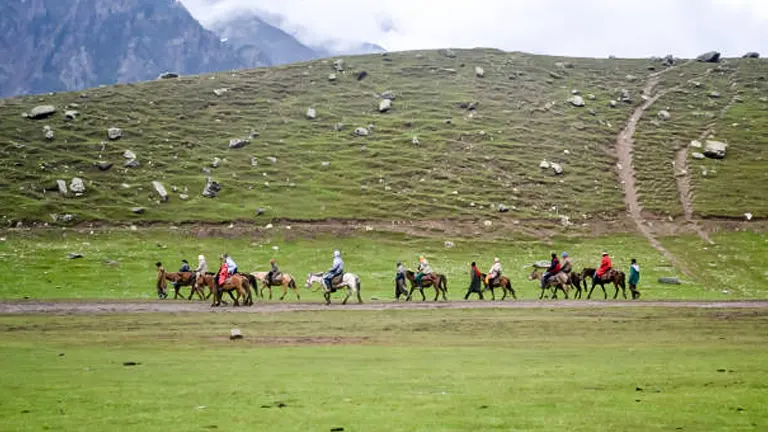 A group of horseback riders trekking across a green, grassy valley with a trail leading up a hill, set against a backdrop of mountainous terrain partially shrouded by mist.
