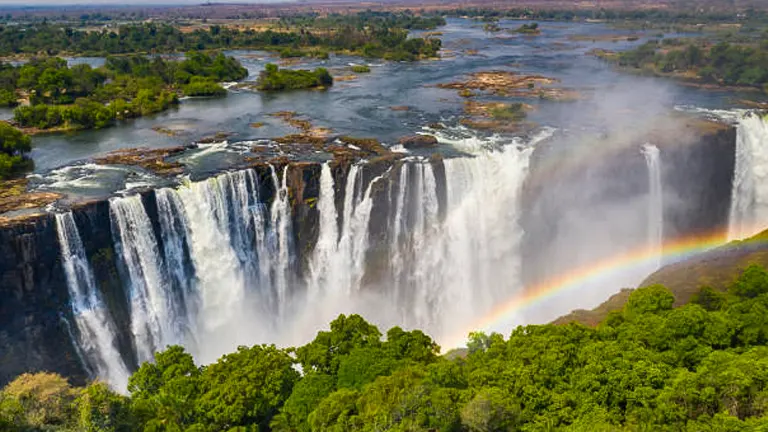 Aerial view of Victoria Falls with a rainbow arching over the misty chasm, surrounded by the lush greenery of the African wilderness and the tranquil Zambezi River upstream.