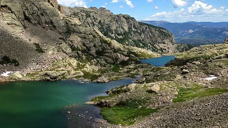 High-altitude lakes nestled among rugged mountain slopes, with green vegetation contrasting with the stone and sparse snow patches, under a partly cloudy sky.