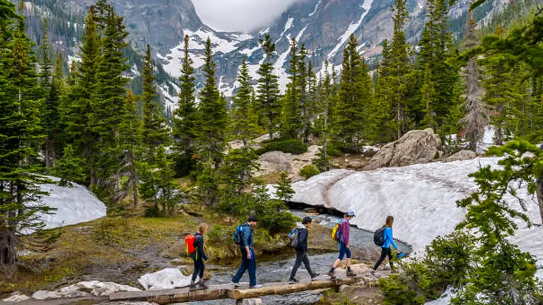 A group of hikers crossing a wooden bridge over a stream in a mountainous area with lingering snow patches, pine trees, and cloudy skies overhead.