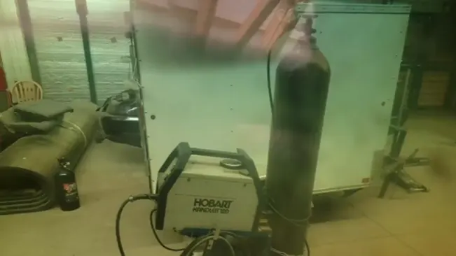 Alt text: "A welding setup with a Hobart Handler 210 welding machine, gas cylinder, and other equipment in a workshop environment, seen through a greenish tint, possibly due to a welding screen.