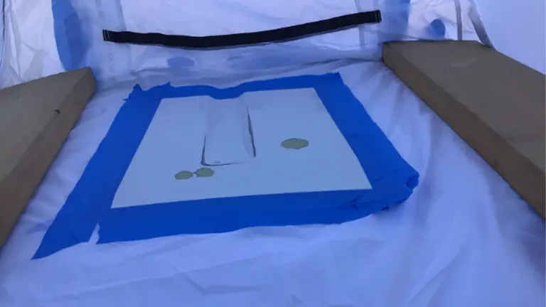 Inside a spray shelter, a flat item is taped off with blue painter's tape for a precise paint job, surrounded by wooden boards.