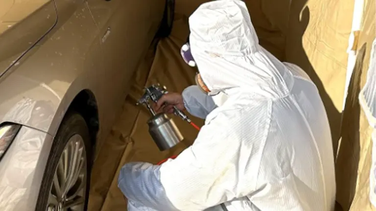 A person in protective coveralls and a mask using a spray gun to paint a car inside a tan paint booth.