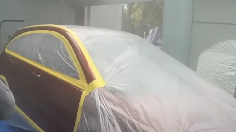 A car prepared for painting, wrapped in translucent plastic sheeting with yellow tape outlining the windows, inside a bright inflatable paint booth.