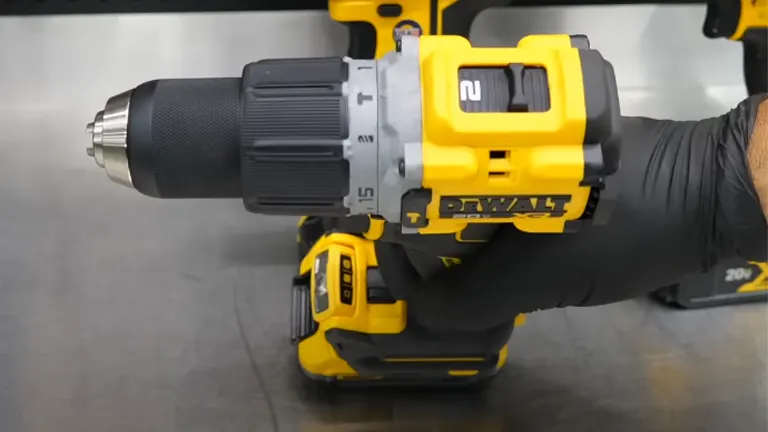 A person's hand wearing a black glove is holding a DEWALT 20V Max XR cordless drill, displaying its gray chuck and yellow-and-black body, set against a background with similar power tools.
