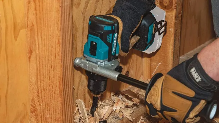 Hands wearing protective gloves are using a Makita cordless hammer drill to bore into a wooden beam, with wood shavings indicating active drilling work.