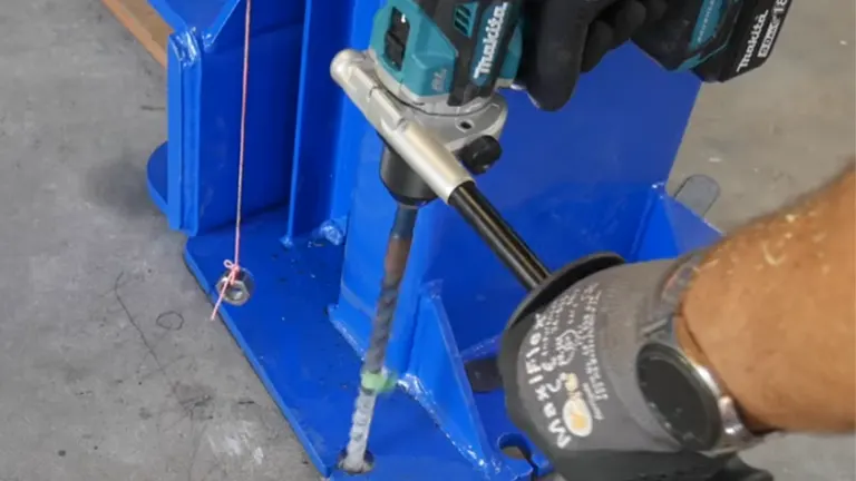 A close-up of a Makita cordless hammer drill being used to drill into concrete, with focus on the tool and drill bit, and the user's gloved hands guiding the process.