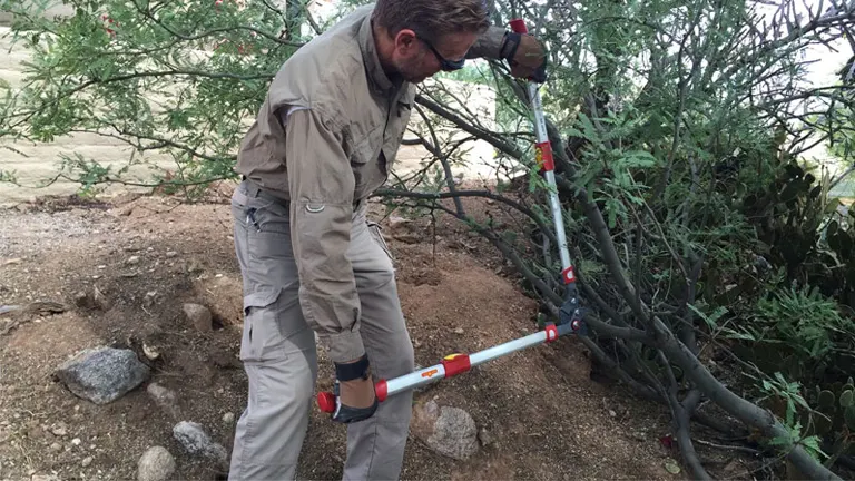 A man in outdoor work attire using long-handled, red and silver bypass loppers to prune a shrub in a rocky terrain garden.