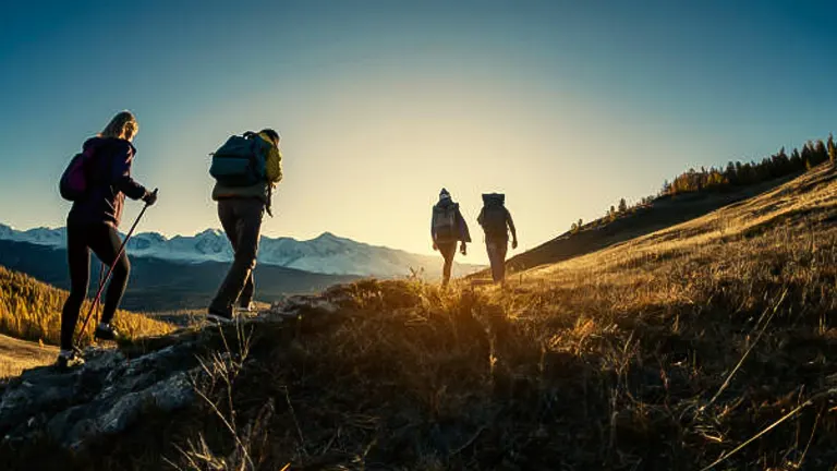 Group of hikers trekking on a mountain trail at sunset, with the golden hour light illuminating the grassy slope and distant mountain peaks.