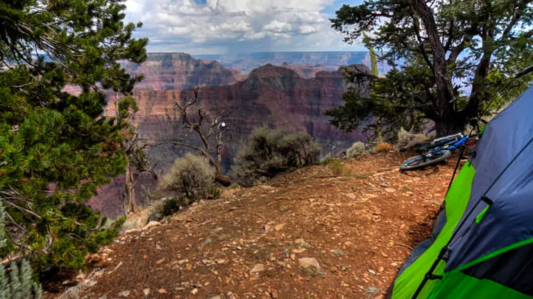 A green tent is pitched on the edge of a cliff with a spectacular view of the Grand Canyon, while a mountain bike leans against a nearby tree, suggesting an adventurous camping trip.
