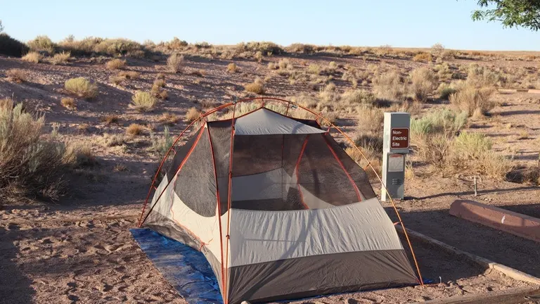 A tent is pitched on a sandy campsite in a desert area, marked by a "Non-Electric Site" sign, indicating a primitive camping setting with a backdrop of sparse desert vegetation under a clear sky.