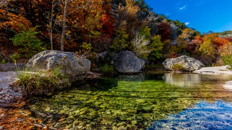 A crystal-clear creek with large boulders and lush autumn foliage in a forested area under a bright blue sky.

