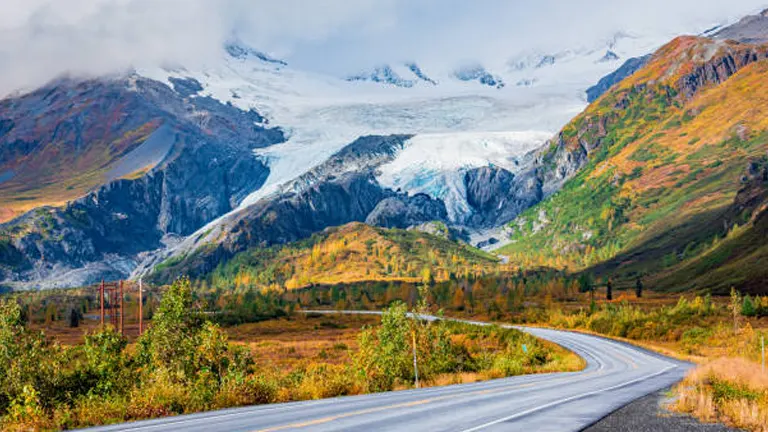 A winding asphalt road meandering through a vibrant Alaskan valley with autumn colors, leading towards a majestic glacier nestled between mountain peaks under a soft-lit sky.