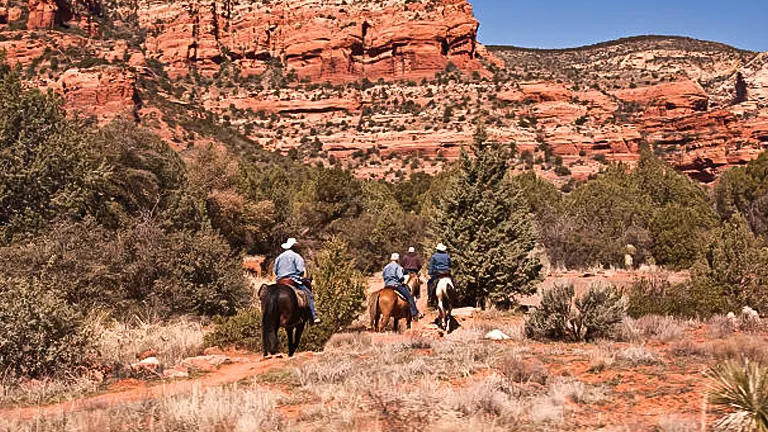 Three riders on horseback traversing a desert trail with striking red rock formations in the background under a clear blue sky.

