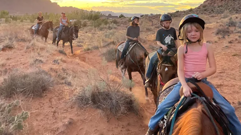 A group of horseback riders, including children, on a trail at sunset, with the warm glow of the sky complementing the red desert terrain.