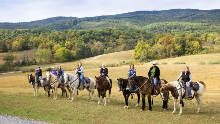 A group of contented horseback riders on a gravel path, with a backdrop of rolling autumnal hills and a cloudy sky, creating a serene countryside tableau.
