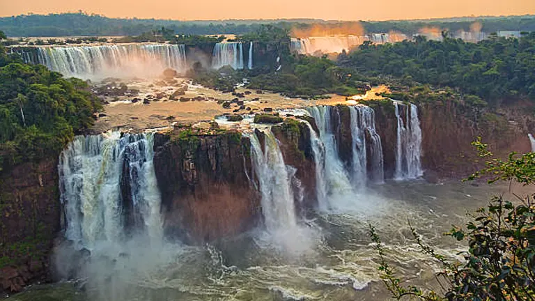 Panoramic view of Iguazu Falls at sunset, with golden light bathing the expansive cascade network and surrounding rainforest, as mist rises from the tumultuous river below.