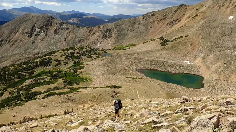 A lone hiker trekking down a rocky mountain path towards a small, secluded teal lake in a vast, open alpine landscape under a cloudy sky.
