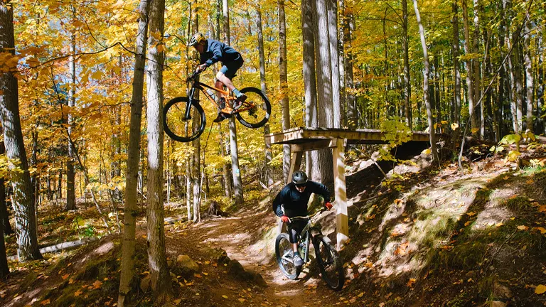 A mountain biker catches air over a jump on a forest trail, while another rider cycles below on a sunny autumn day, surrounded by trees with golden foliage.
