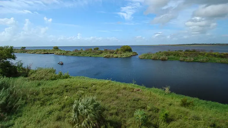 A panoramic view of a serene wetland with scattered green islets, a small boat in the water, and lush vegetation under a vast, cloudy sky.

