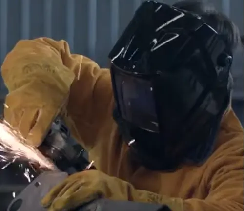 Welder in protective gear using a grinding tool, with a welding helmet on.